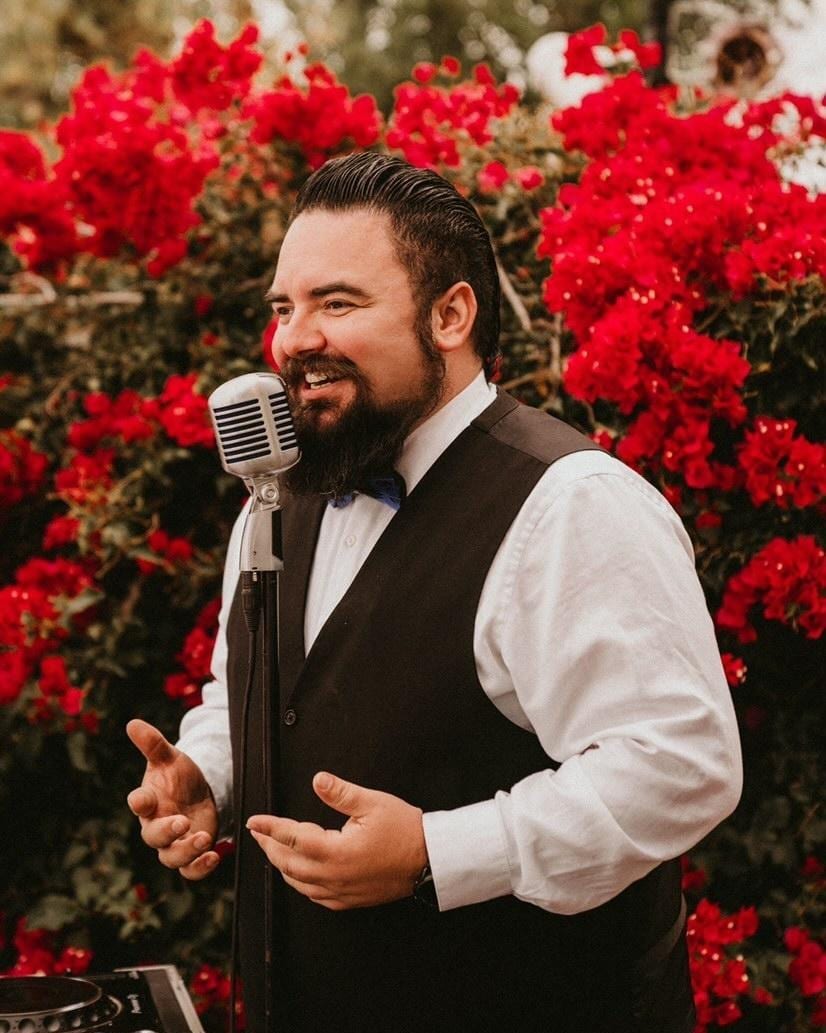 Man in formal attire speaking into a vintage microphone with vibrant red flowers behind.